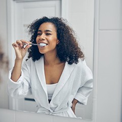 woman in a white bathrobe brushing her teeth in front of a mirror 