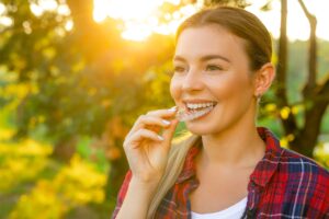 Woman wearing red plaid in front of sunny trees inserting top Invisalign tray