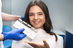 Woman with brown hair in dental chair smiling and gesturing to a shade guide being held near her face