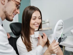 Dentist holding veneers while woman smiles into mirror.