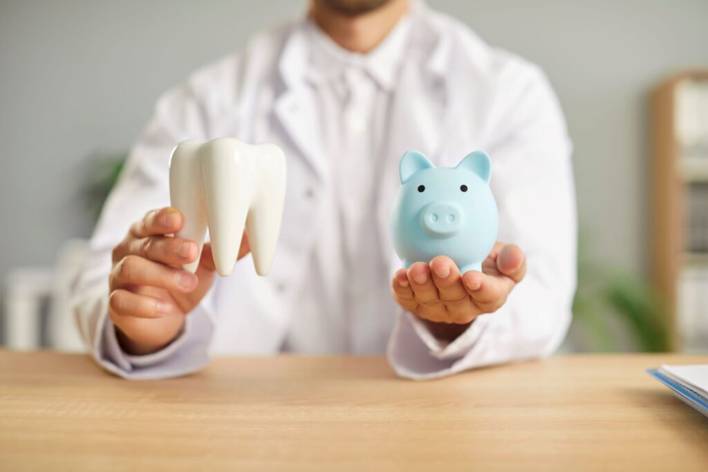 Dentist in white jacket holding a large model tooth and small blue piggy bank